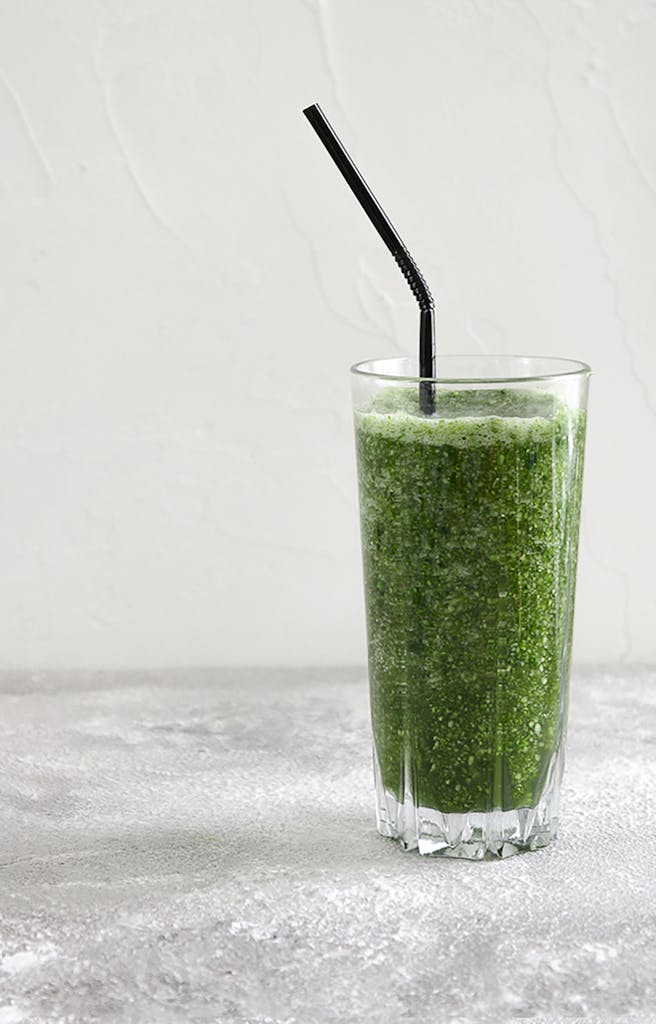 A healthy, refreshing green smoothie served in a tall glass with a black straw, against a minimalist background.