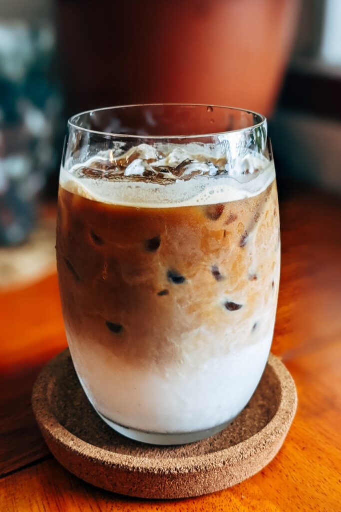A close-up of a delicious iced coffee in a glass with a cork coaster on a wooden tabletop.