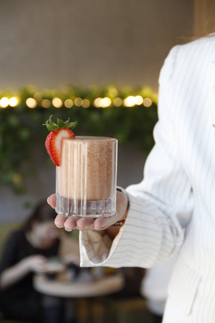 A woman in a white suit holds a creamy strawberry smoothie garnished with fresh fruit in a café setting.